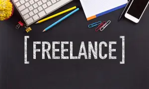 Biurko z klawiaturą, telefonem, notatnikiem i kredkami. Słowo "FREELANCE" na czarnym tle. To jest właśnie freelancer co to.