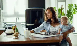 Młoda mama z dzieckiem na rękach pracuje przy laptopie. Czy na macierzyńskim można pracować? Tak, wiele mam łączy opiekę z pracą zdalną.