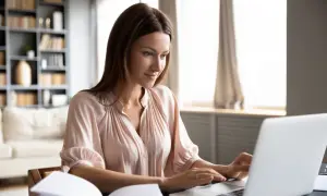 Kobieta pracuje przy laptopie, realizując chałupnictwo z dowozem do domu. W tle regał z książkami i kanapa.