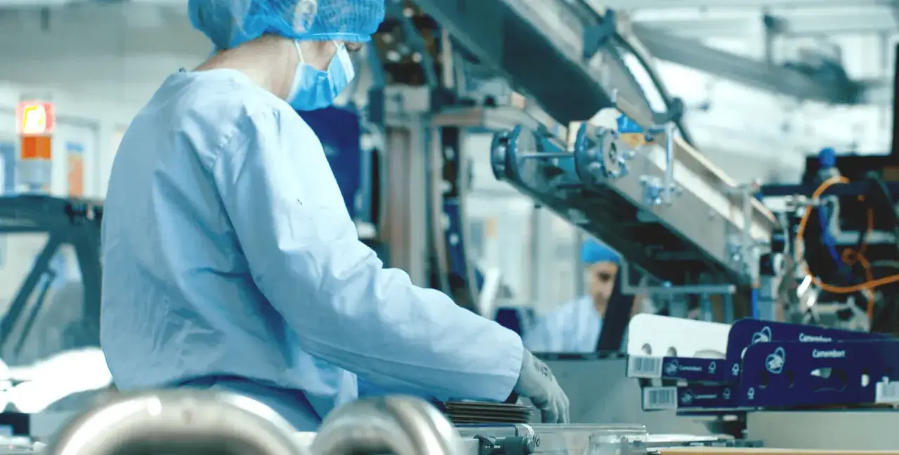 Pracownik w fartuchu i czepku pracuje przy linii produkcyjnej, pakując sery. Stanowiska pracy w mleczarni są zautomatyzowane.