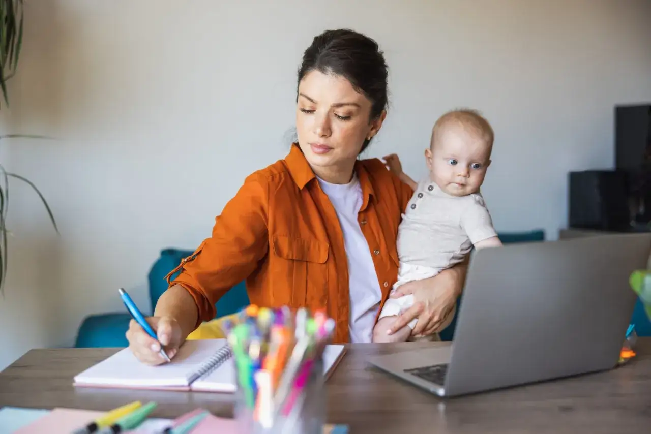 Kobieta z dzieckiem na rękach pracuje przy laptopie, notując coś w zeszycie. Czy na macierzyńskim można pracować? Tak, jak widać.