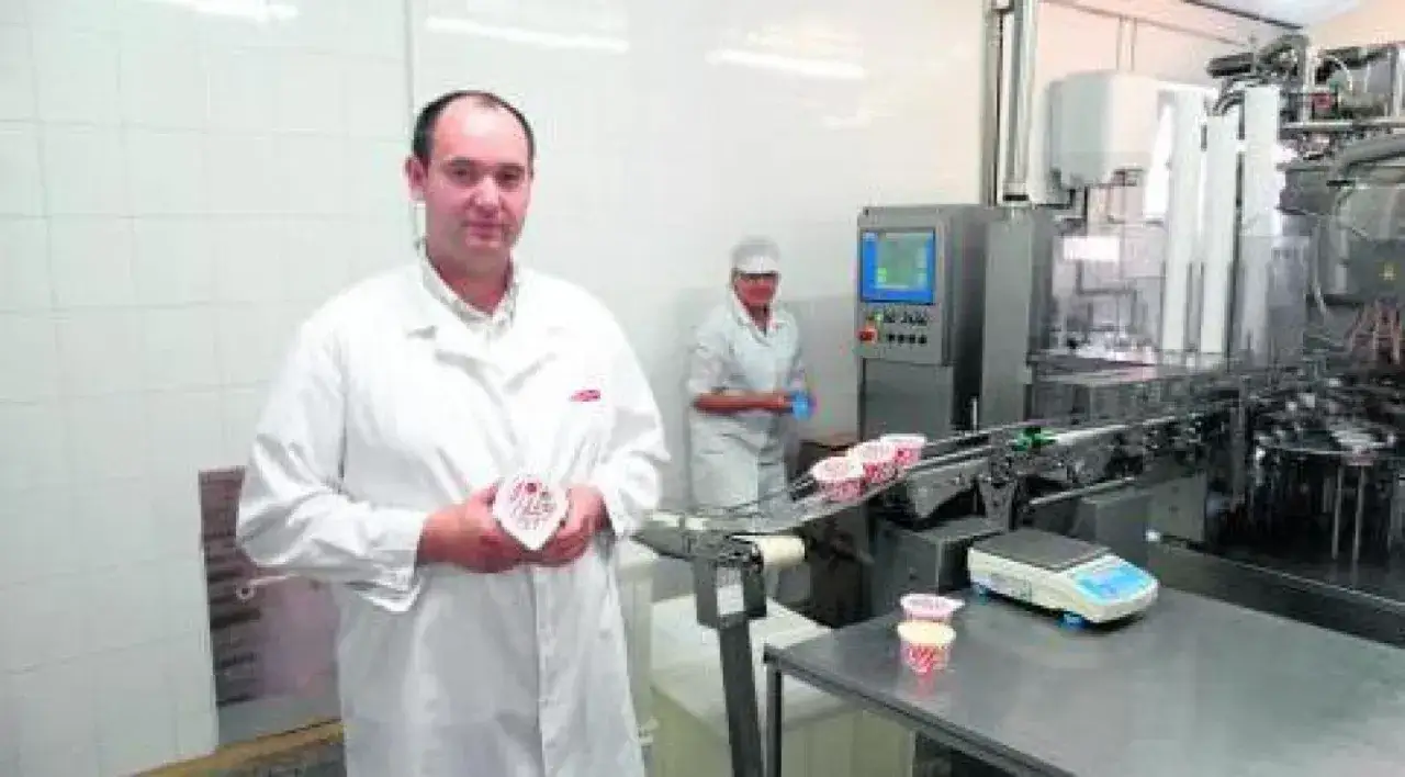Pracownik w białej kurtce prezentuje jogurt na tle linii produkcyjnej w mleczarni. W tle widać pracownicę i maszyny.