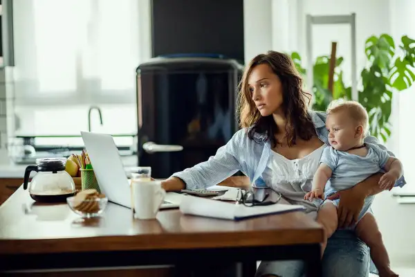 Młoda mama z dzieckiem na rękach pracuje przy laptopie. Czy na macierzyńskim można pracować? Tak, wiele mam łączy opiekę z pracą zdalną.
